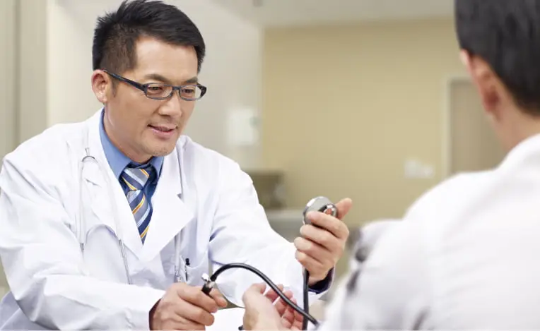 白衣を着て眼鏡をかけた男性医師が、患者の腕に聴診器を当てながら優しく微笑んでいる。患者の背中が一部写っている。