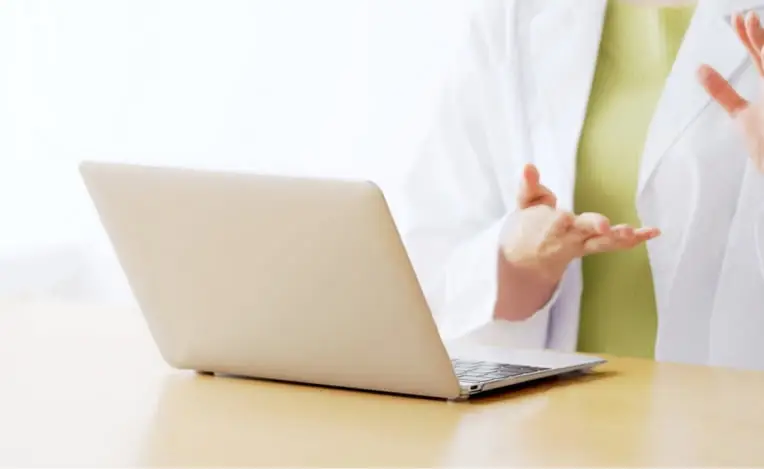 白衣を着た医療従事者がノートパソコンの前で手を使って説明している様子
